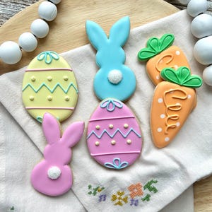 May include: Easter-themed sugar cookies in the shapes of eggs, bunnies, and carrots. The cookies are decorated with pastel colors, including yellow, pink, and blue, and feature intricate icing details. A white cloth with floral embroidery is underneath the cookies.