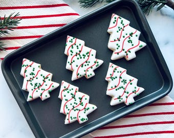 Puede incluir: Una bandeja para hornear de color gris oscuro contiene cinco galletas con forma de árbol de Navidad. Las galletas son blancas con líneas de glaseado rojo y chispas verdes. Un paño a rayas rojas y blancas y ramitas de hoja perenne están en el fondo.