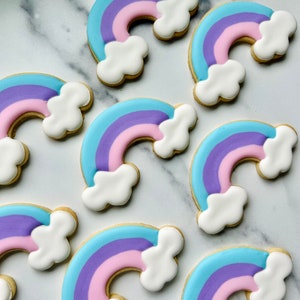 May include: A collection of rainbow-shaped sugar cookies decorated with white icing clouds. The cookies are a light blue, pink, and purple color.
