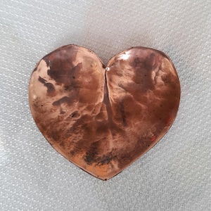 May include: A heart-shaped copper dish with a textured surface. The dish has a warm, reddish-brown color with darker areas, and a slight indentation in the center. The heart shape is clearly defined, and the overall appearance is rustic and handcrafted.