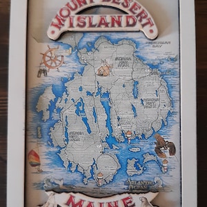 May include: A framed map of Mount Desert Island, Maine, with a white border. The map features blue water and a detailed depiction of the island's geography, with the text "Mount Desert Island" at the top and "Maine" at the bottom.