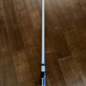 K&ouml;nnte beinhalten: Ein Lichtschwert-Spielzeug mit einer langen, zylindrischen wei&szlig;en Klinge, die an einem Griff aus silbernem und schwarzem Metall befestigt ist. Der Griff verf&uuml;gt &uuml;ber einen strukturierten Bereich, ein seitliches Bedienfeld und einen glatten silbernen Emitter.