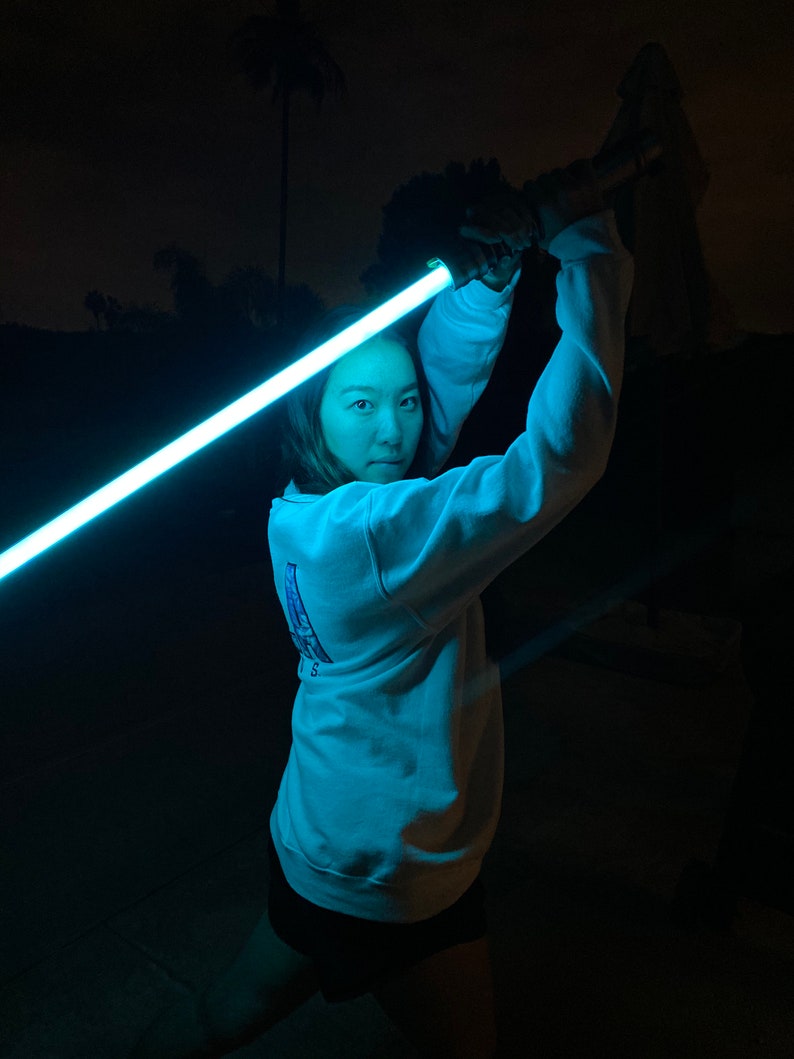 以下が含まれることがあります： 暗い場所で青いライトセーバーを持っている人。その人は白いスウェットシャツと黒いショートパンツを履いています。