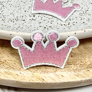 Peut inclure: Cinq patchs brodés en forme de couronne rose et blanche sur une assiette blanche et une surface en bois.