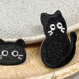 Peut inclure: Trois patchs de chat en feutre noir avec des yeux et des moustaches brodés en blanc. Les chats sont dans des poses différentes, l'un assis, l'autre debout et le dernier regardant droit devant.