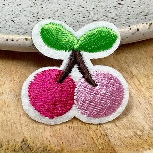 Peut inclure: Un patch en feutre blanc avec un motif brodé de deux cerises roses avec des feuilles vertes et des tiges brunes.