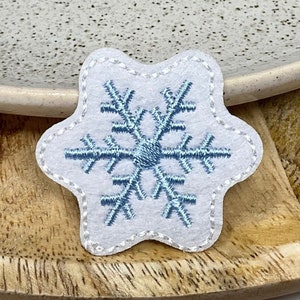 May include: A white felt snowflake applique with blue embroidery. The snowflake is a six-pointed star with a circle in the centre.