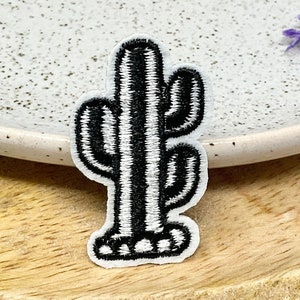Peut inclure: Un écusson brodé représentant un cactus en noir et blanc. Le patch présente un motif détaillé de cactus sur fond blanc avec des coutures noires. Il est posé sur un plateau en bois, à côté d'une assiette blanche mouchetée.