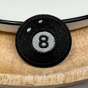 Könnte beinhalten: Schwarzer, gestickter Aufnäher mit der Zahl 8 in einem weißen Kreis, der einer Billardkugel ähnelt. Der runde Aufnäher hat einen schwarzen Rand und liegt auf einer Holzoberfläche und einem weißen Teller.