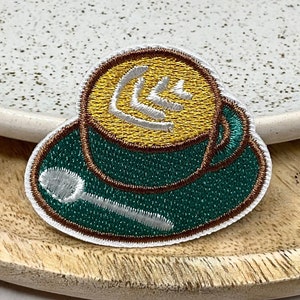 May include: An embroidered patch depicting a coffee cup with a white spoon. The cup is green and brown with a yellow interior and a white design on top.