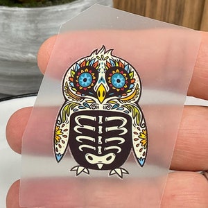 Peut inclure: Un autocollant transparent avec un motif de hibou crâne de sucre coloré. Le hibou a des yeux bleus, un bec jaune et un motif de squelette noir et blanc.