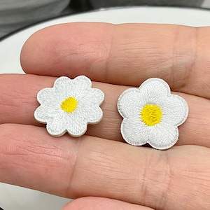 Könnte beinhalten: Zwei weiße Gänseblümchen-Blumenaufnäher mit gelben Zentren werden gezeigt. Die Aufnäher sind mit weißem Faden bestickt und haben ein detailliertes Blütenblattdesign. Die Aufnäher werden in einer Hand gehalten und liegen auf einem weißen Teller.