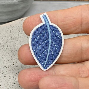 May include: Three blue embroidered fabric leaf patches with white outlines. The leaves are on a white surface.
