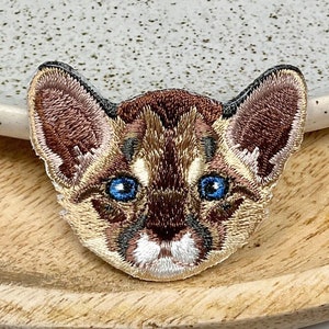 Può includere: Toppa ricamata con il viso di un gatto con pelliccia marrone e occhi blu. La toppa è su una superficie di legno.