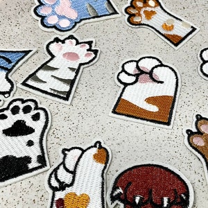 Peut inclure: Une collection de patchs brodés en forme de pattes de chat, de différentes couleurs et motifs, dont blanc, marron et jaune. Les patchs sont disposés sur une assiette en céramique mouchetée.