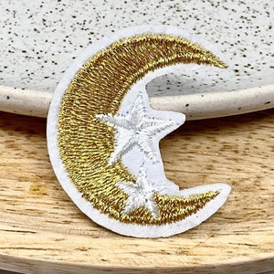 May include: A gold crescent moon with two white stars embroidered on a white fabric patch.