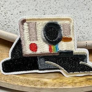 Peut inclure: Patch brodé thermocollant représentant un appareil photo instantané vintage avec une bande arc-en-ciel et un point rouge. L'appareil photo est beige avec un objectif noir et une base noire.