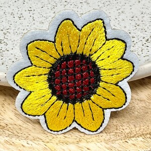 Peut inclure: Un patch brodé de tournesol jaune avec un centre rouge. Le patch est sur un fond blanc.