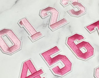 May include: Pink felt iron-on numbers 0 through 9. The numbers are in a varsity style font with a white outline.