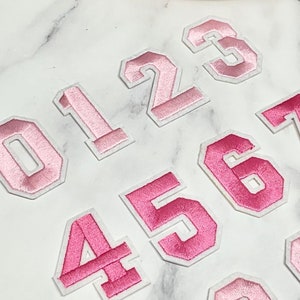 Pode incluir: Números de feltro rosa para passar a ferro de 0 a 9. Os números estão em um estilo de fonte universitário com contorno branco.