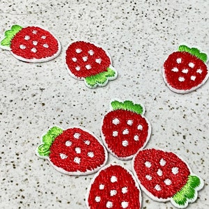 May include: Seven red embroidered strawberry patches with white seeds and green leaves on a white speckled surface.