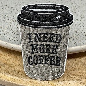 May include: An embroidered patch featuring a grey coffee cup with the text "I NEED MORE COFFEE" in black.