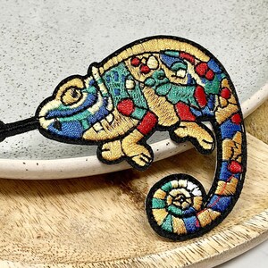 Peut inclure: Un patch brodé de caméléon coloré avec un motif jaune, rouge, bleu et vert. Le patch est sur une surface en bois.