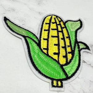 Pode incluir: Um remendo bordado para passar a ferro de uma espiga de milho amarela com folhas verdes.