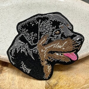 Peut inclure: Un patch brodé représentant la tête d'un Rottweiler, en noir et marron. Le chien regarde vers la droite et a la langue qui pend.