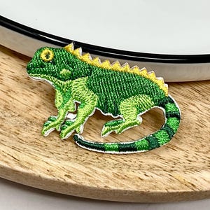 Peut inclure: Un écusson brodé représentant un iguane vert avec une crête jaune le long du dos et une longue queue rayée. Le lézard est représenté de profil avec des coutures détaillées montrant sa peau texturée, ses membres et son œil.