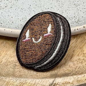 May include: A brown and black embroidered patch of a smiling cookie with a white face and pink cheeks.