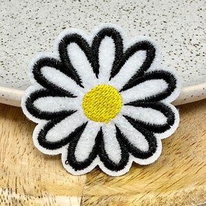 Könnte beinhalten: Weißer gestickter Gänseblümchen-Aufnäher mit schwarzem Rand und gelber Mitte. Der Aufnäher befindet sich auf einer Holzoberfläche.