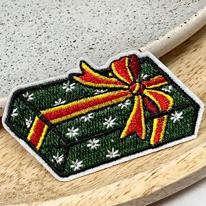 Peut inclure: Boîte cadeau verte et blanche avec un nœud rouge et jaune. La boîte cadeau est brodée de flocons de neige blancs.