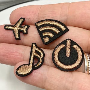 Peut inclure: Un ensemble de cinq petits écussons brodés. Les écussons comprennent un avion doré, un symbole WiFi, une note de musique et un bouton d'alimentation, tous avec des coutures dorées sur fond noir. Les écussons sont présentés sur une assiette blanche.