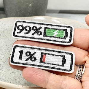Peut inclure: Deux patchs thermocollants brodés blancs avec des contours noirs. Un patch montre une icône de batterie avec une barre verte et le texte "99%". L'autre patch montre une icône de batterie avec une barre rouge et le texte "1%".