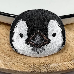 May include: Circular embroidered patch depicting a penguin's face. The patch is black and white with detailed stitching, showing the penguin's eyes, beak, and facial features. The patch is on a wooden surface, likely for application to clothing or accessories.