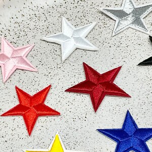 Peut inclure: Douze étoiles en feutre brodées de différentes couleurs, dont le rouge, le bleu, le vert, le jaune, le rose, le violet et le blanc. Les étoiles sont disposées en cercle sur une assiette blanche.