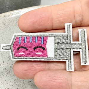 Peut inclure: Un patch brodé thermocollant gris et rose représentant une seringue avec un visage souriant. La seringue est remplie de liquide rose et comporte des marques bleues.