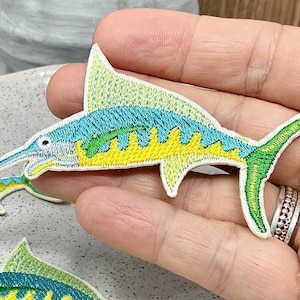 May include: Embroidered patches featuring a marlin fish design. The patches have a white background with a detailed depiction of the fish in shades of blue, yellow, and green. The patches are on a speckled ceramic plate.