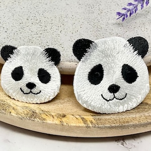 Peut inclure: Deux visages de panda brodés en blanc avec des yeux et des nez noirs sur une surface en bois.