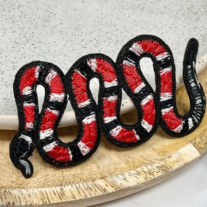 May include: An embroidered snake patch in red, black and white. The snake is curled up in a coil shape.
