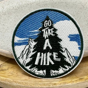 May include: A round, white embroidered patch with a green pine tree and the text "Go Take A Hike" in white. The background is a blue sky with white clouds.