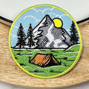 May include: Embroidered patch depicting a mountain scene, with a blue sky, white clouds, a yellow sun, green trees, a grey mountain, and an orange tent.