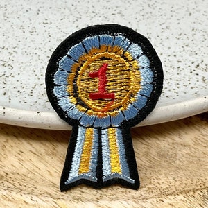Peut inclure: Un ruban brodé bleu et or avec un cercle doré et le chiffre "1" en rouge. Le ruban est sur une surface en bois.
