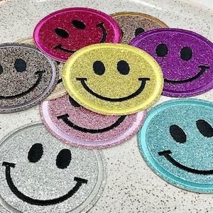 Peut inclure: Un ensemble de sept patchs smiley en paillettes de différentes couleurs, dont le rose, le jaune, le marron, le violet, le bleu sarcelle et l'argent. Les patchs sont disposés sur une assiette blanche à surface mouchetée.