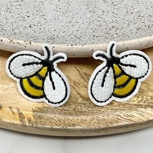 Può includere: Due toppe ricamate a forma di ape con strisce gialle e nere e ali bianche. Le toppe sono su una superficie di legno.