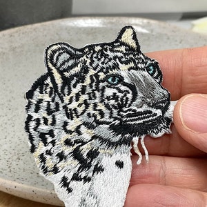 May include: Embroidered patch featuring a snow leopard's head with detailed stitching. The patch showcases a black, white, and gray color palette with striking blue eyes. The design is a detailed representation of the animal's features, suitable for clothing or accessories.