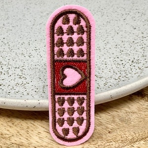 Peut inclure: Un patch en tissu rose en forme de pansement avec un cœur brodé marron au centre. Le pansement a une bordure brodée marron et des détails brodés marron en haut et en bas.