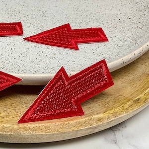 May include: Four red felt arrow patches on a white plate and a wooden surface. The arrows are pointing to the right.