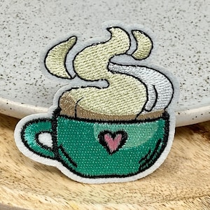 May include: Green coffee cup with a heart and steam coming out of it. The cup is embroidered on a white fabric.